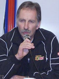 Eine Person spricht in ein Mikrofon und trägt eine Sportjacke mit einem Logo. Im Hintergrund sind farbige Flaggen zu sehen.