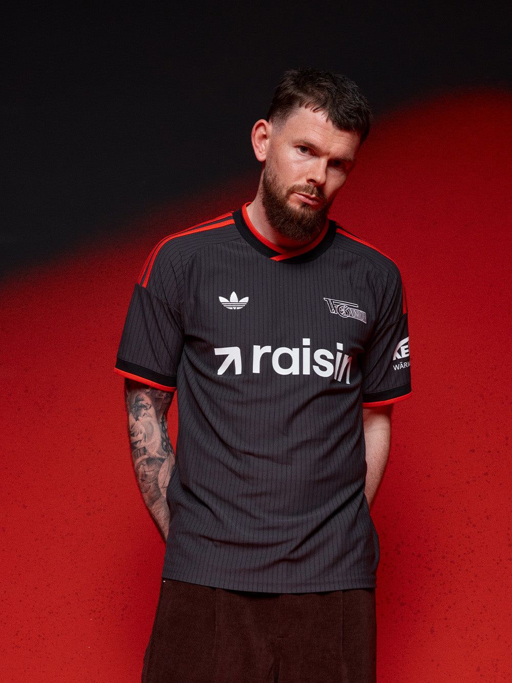 Mann in einem grauen, gestreiften Fußballtrikot mit roten Akzenten steht vor einem roten Hintergrund.