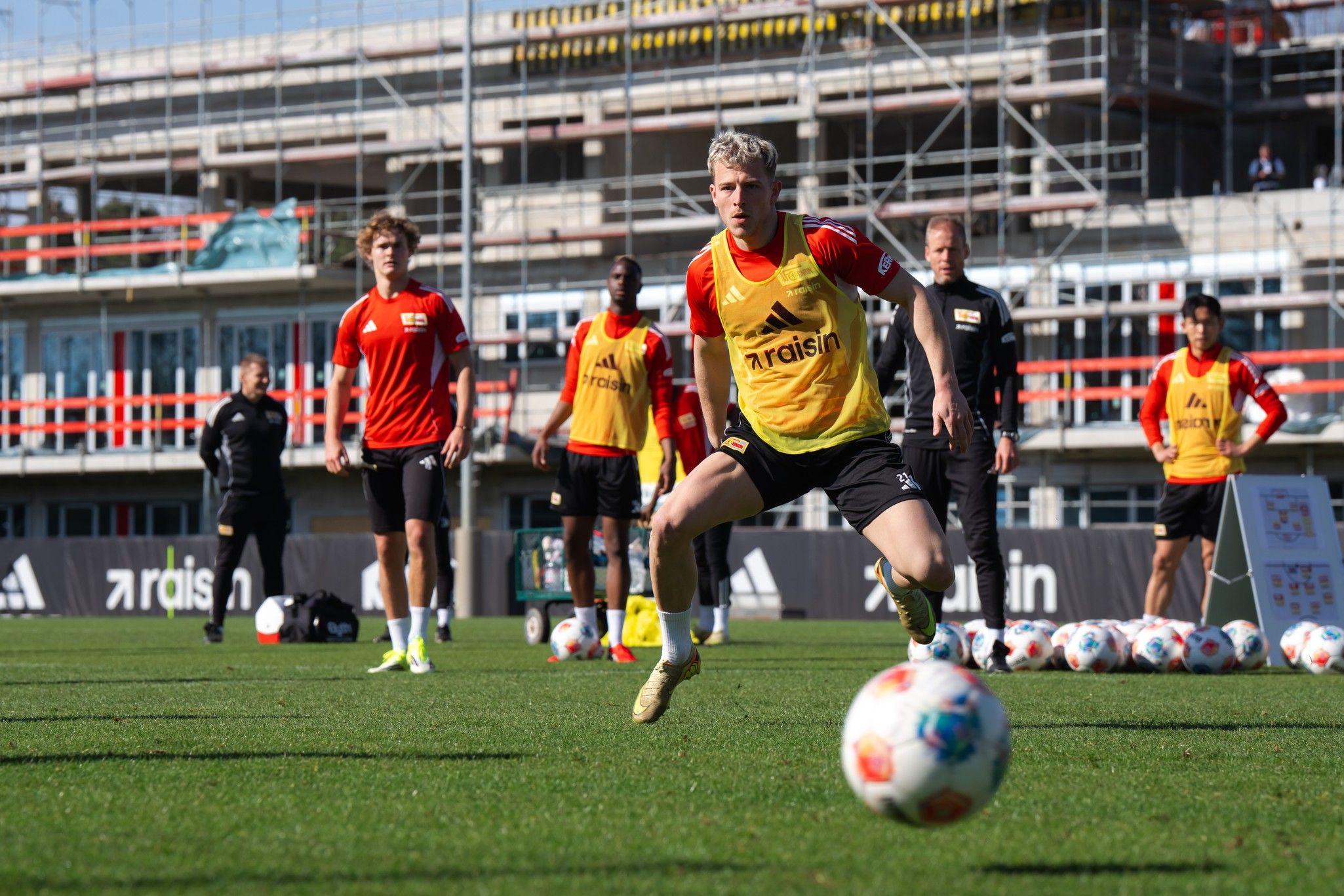 Spieler in gelben Trikots trainieren auf einem Fußballplatz, während andere in roten Trikots zuschauen. Baustelle im Hintergrund.