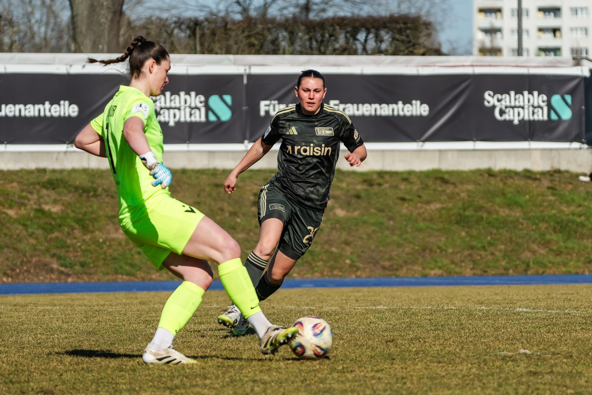 Spielerinnen auf einem Fußballfeld, eine Torhüterin in grüner Ausrüstung und eine Angreiferin in schwarzem Trikot.