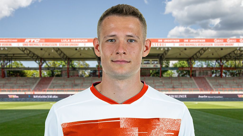 A young soccer player poses in a jersey in front of an empty stadium with a sunny sky in the background.