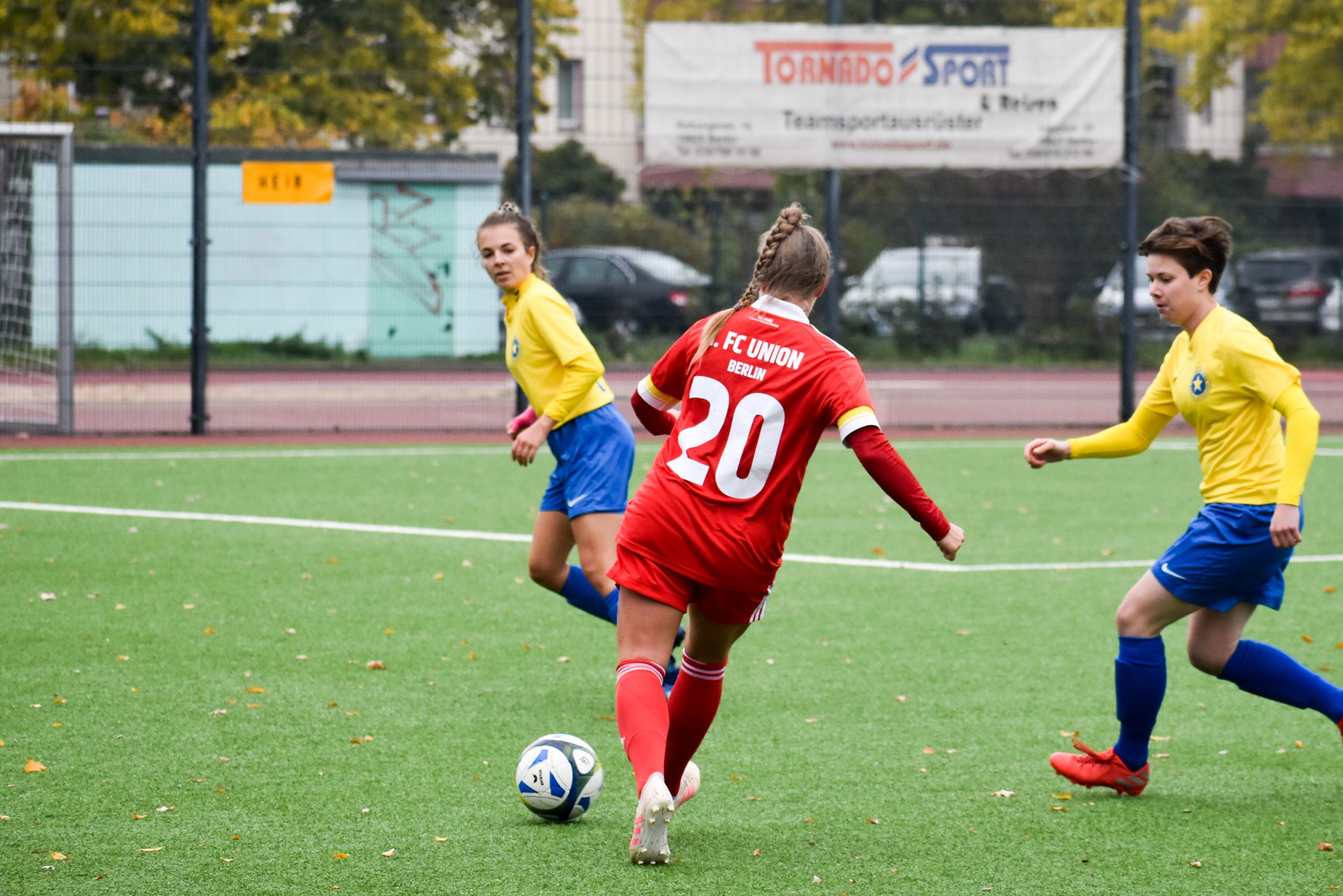 Ein Mädchen in rotem Trikot dribbelt den Ball, während zwei Mädchen in gelben und blauen Trikots ihm folgen.