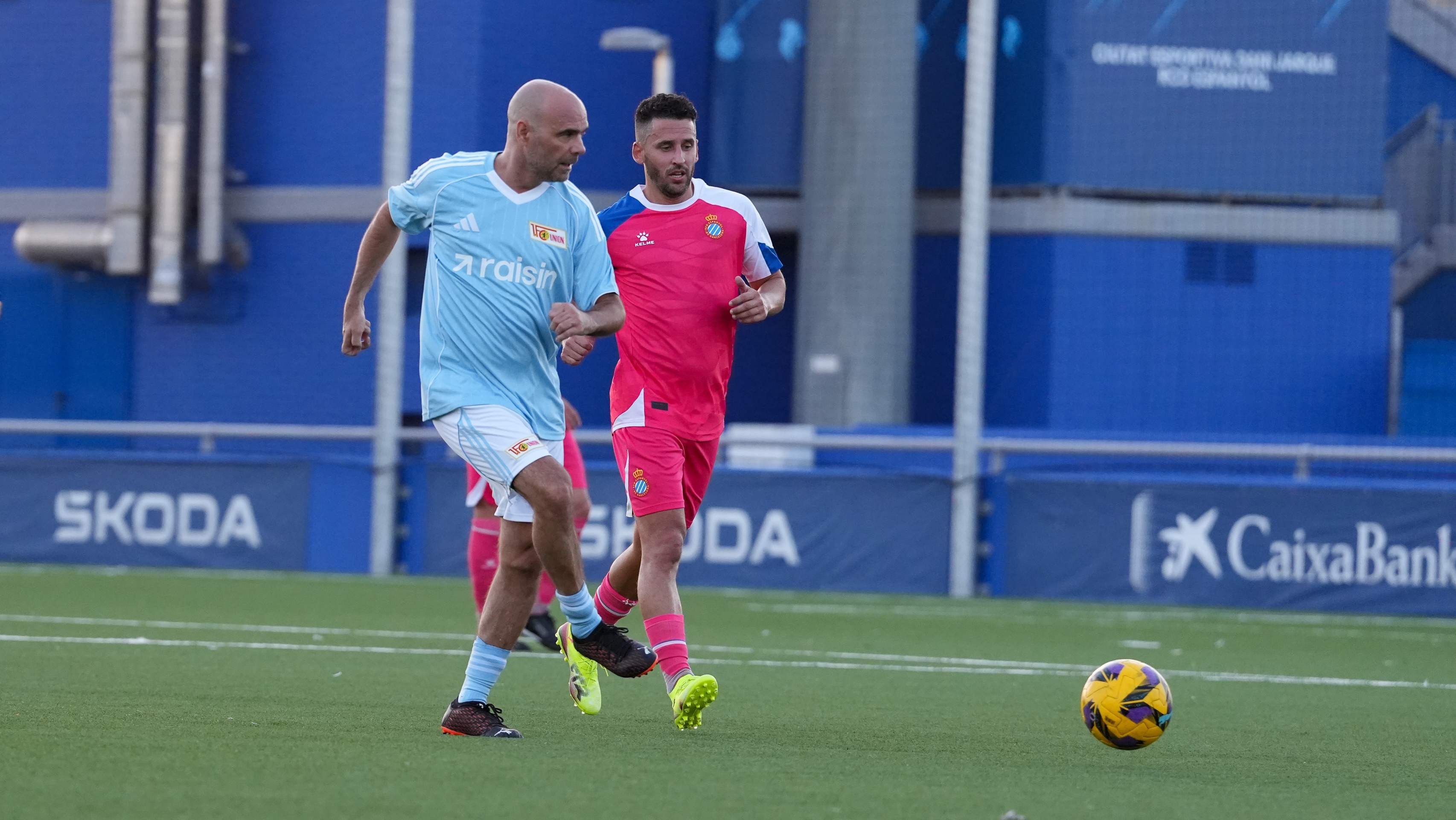 Zwei Männer spielen Fußball. Sie tragen Sportkleidung. Sie sind auf einem grünen Platz.