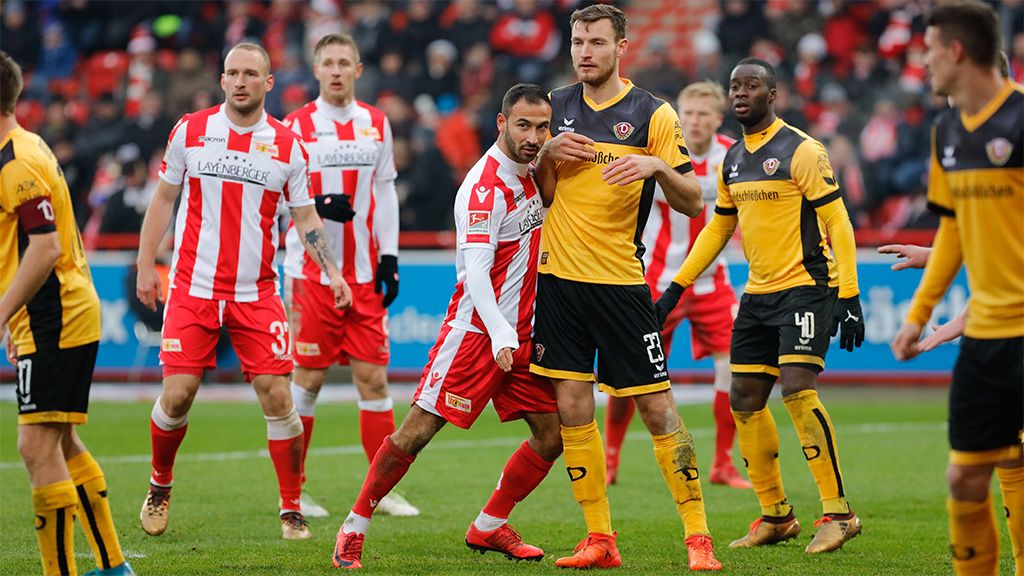 Zwei Fußballspieler stehen auf dem Platz und scheinen sich in einem Wettkampf zu befinden, während andere Spieler im Hintergrund stehen.