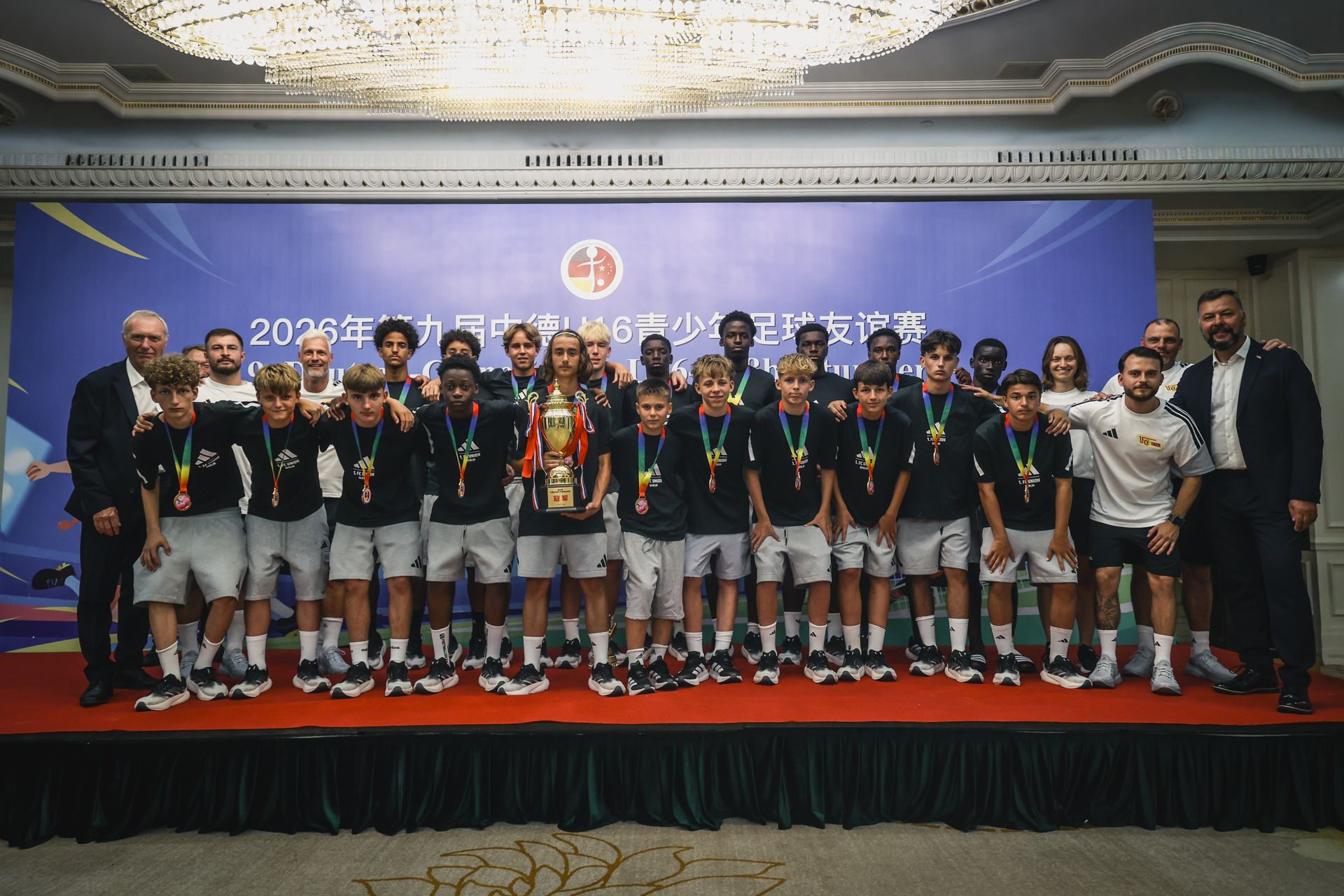 Ein Team aus Jugendlichen in Sportkleidung steht mit Medaillen und einem Pokal auf einer Bühne vor einem Banner.