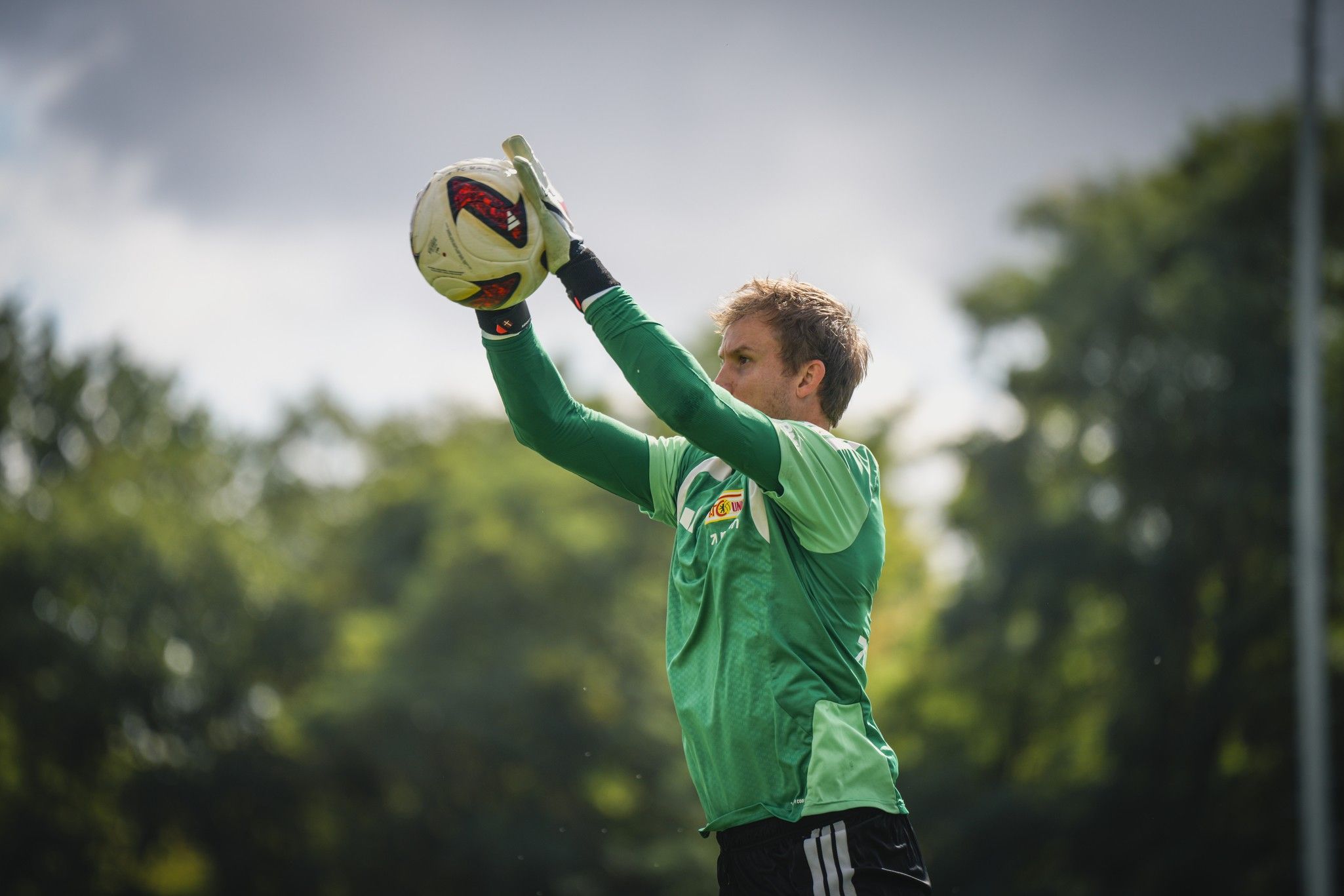 Torwart fängt einen Fußball während des Trainings im Freien. Im Hintergrund Bäume und bewölkter Himmel.