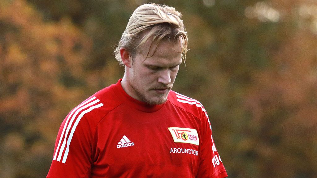 Ein Fußballspieler in einem roten Trikot steht nachdenklich mit gesenktem Kopf auf dem Spielfeld.