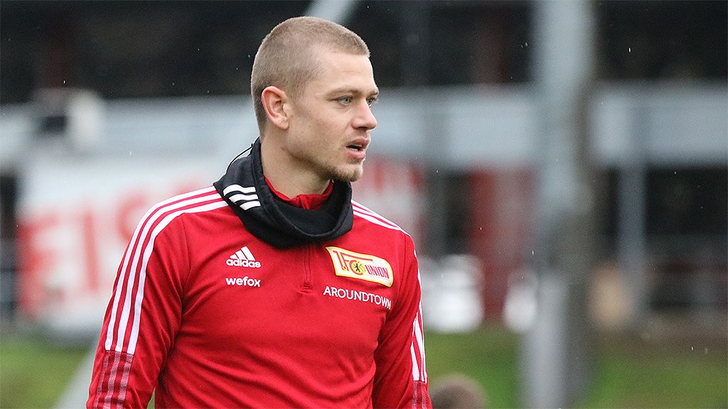 Ein Fußballspieler in roter Trainingskleidung mit einem schwarzen Schal steht auf einem Trainingsplatz.