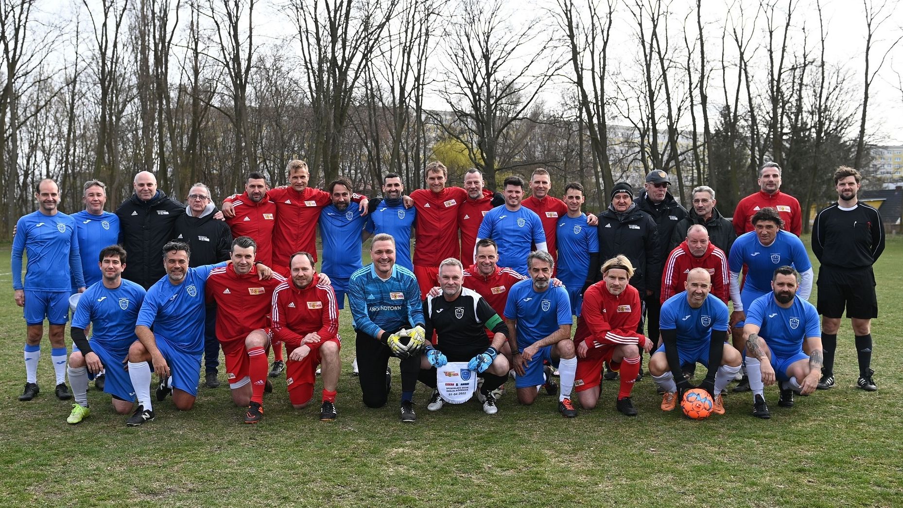 Gruppenfoto von zwei Mannschaften in Sportkleidung auf einem Fußballfeld, umgeben von Bäumen.