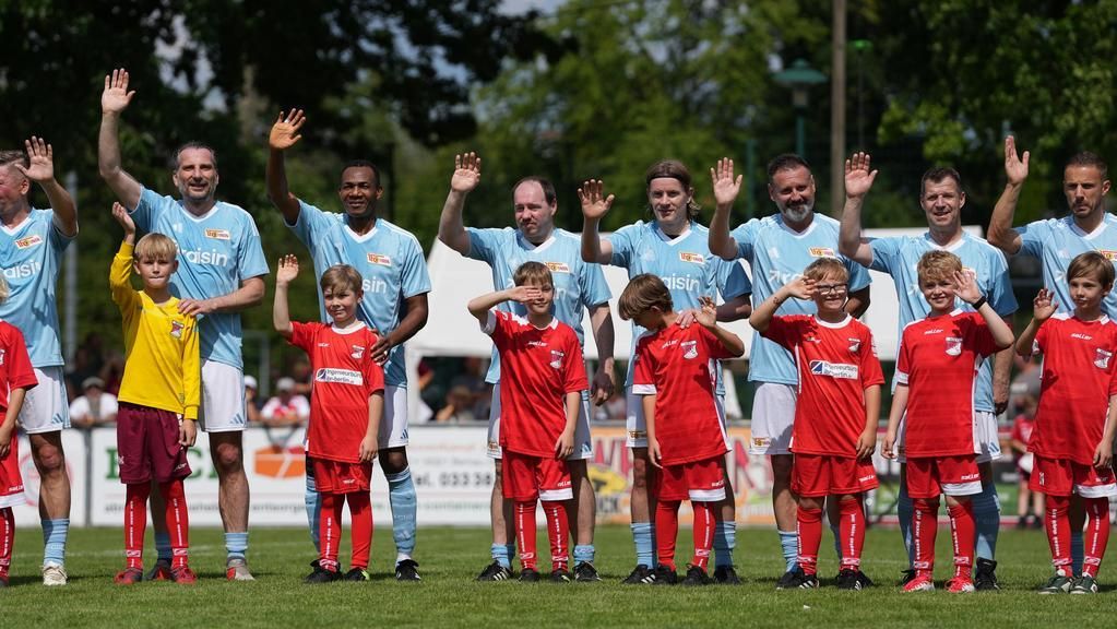 Fünf Fußballspieler in blauen Trikots stehen neben drei Kinderteams in roten Trikots, alle winken in die Kamera.