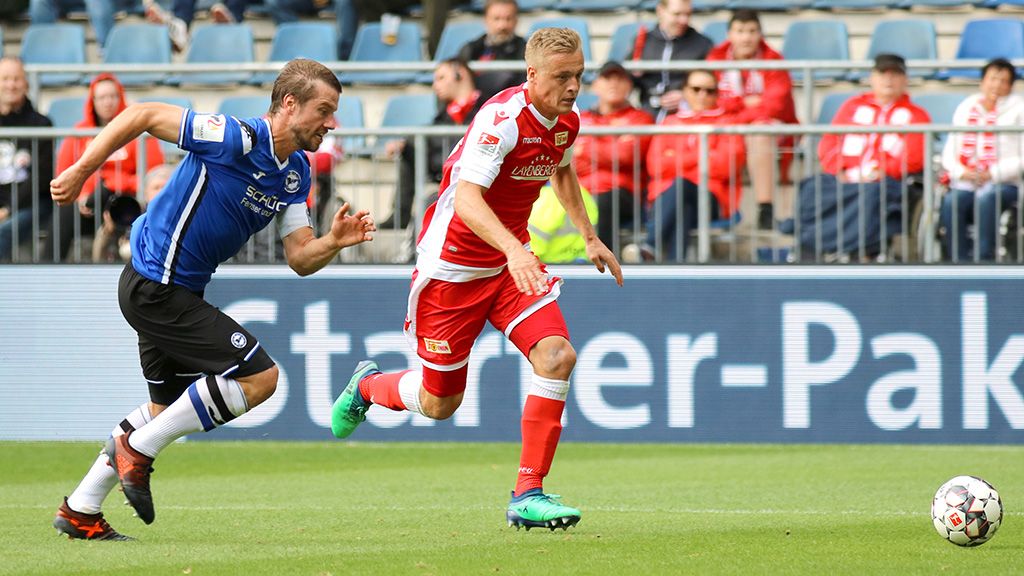 A football player in red sprints with the ball while a player in blue chases him. Spectators are visible in the background.