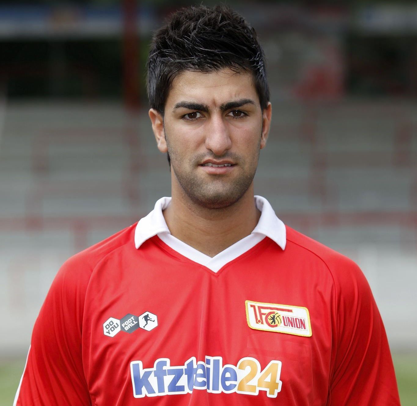Ein junger Mann mit dunklen Haaren trägt ein rotes Fußballtrikot mit dem Logo eines Vereins. Der Hintergrund ist unscharf.