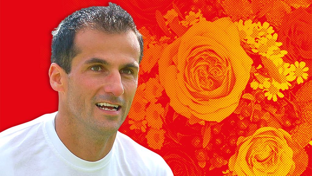 Ein lächelnder Mann in einem weißen T-Shirt vor einem rot-gelben Hintergrund mit großen Rosenmotiven.