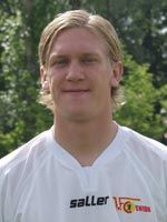 Portait eines lächelnden Mannes in einem weißen Sporttrikot mit Logo, im Hintergrund sind Bäume zu sehen.