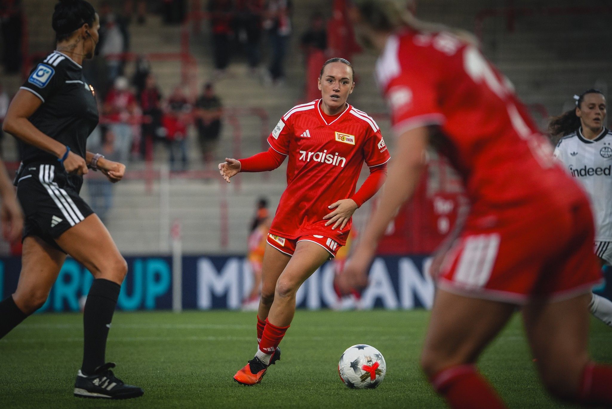 Fußballspiel, Spielerin in rotem Trikot dribbelt den Ball, andere Spielerinnen in Aktion, Stadionhintergrund.