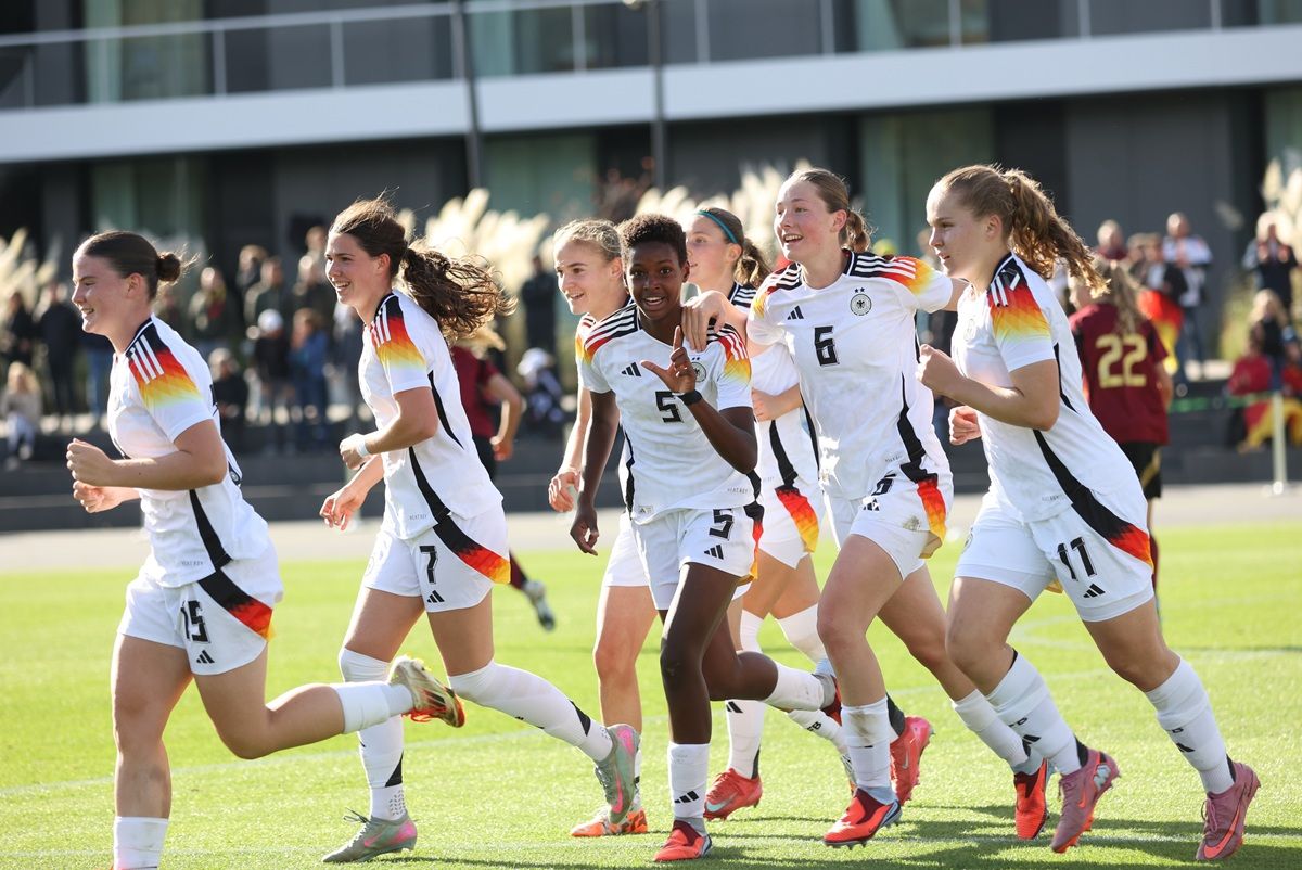Sechs Fußballspielerinnen in weißen Trikots mit Regenbogenstreifen jubeln lachend auf dem Spielfeld.