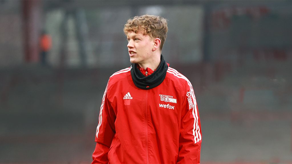 Ein Fußballspieler in roter Trainingsjacke mit Schal steht auf einem Platz und schaut im Hintergrund.