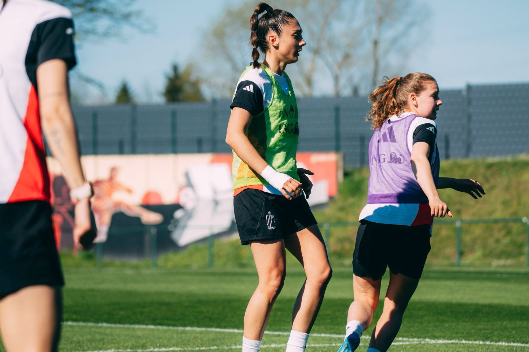 Zwei Fußballspielerinnen beim Training auf dem Spielfeld, eine trägt ein grünes, die andere ein lila Trainingshemd.