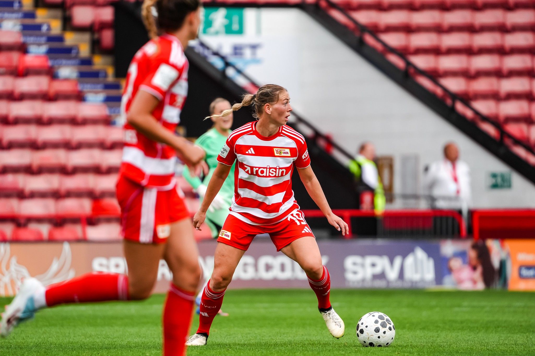 Eine Fußballspielerin in rotem Trikot mit gestreiftem Design spielt auf dem Platz, während eine Mitspielerin ebenfalls in rot zusieht.