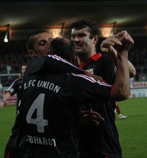 Fußballspieler in schwarzen Trikots feiern einen Torerfolg und umarmen sich auf dem Spielfeld.
