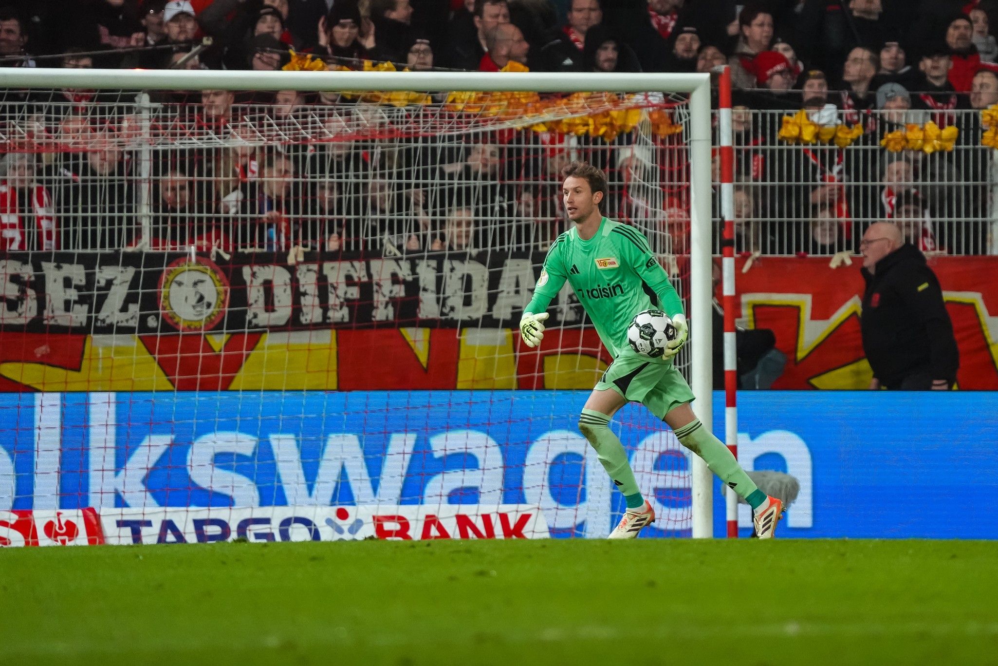 Torwart in grüner Ausrüstung steht im Tor, Fans im Hintergrund mit Bannern und lebhaften Farben.