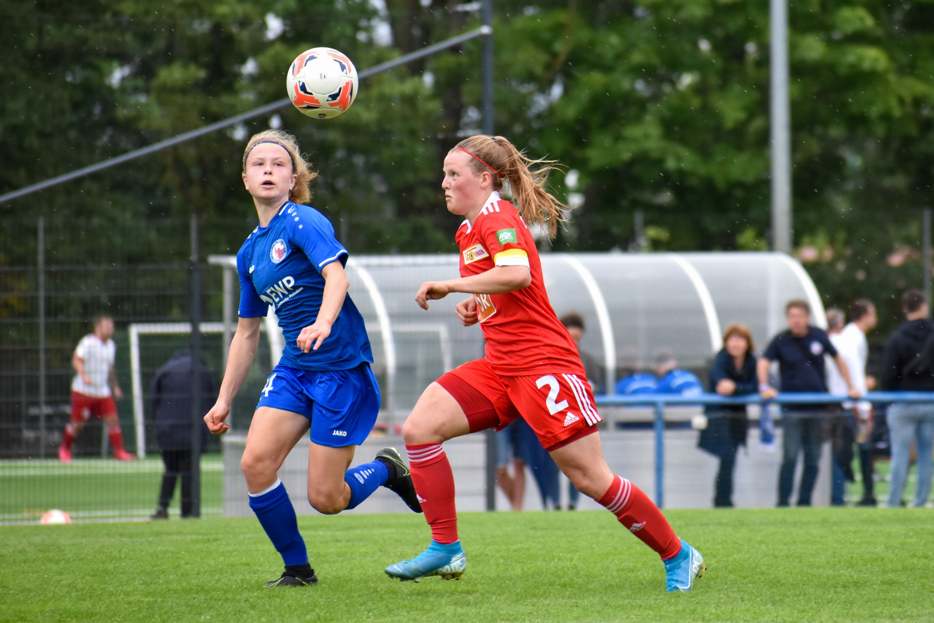 Zwei Fußballspielerinnen in Aktion auf einem Spielfeld, während der Ball in der Luft fliegt. Zuschauer im Hintergrund.