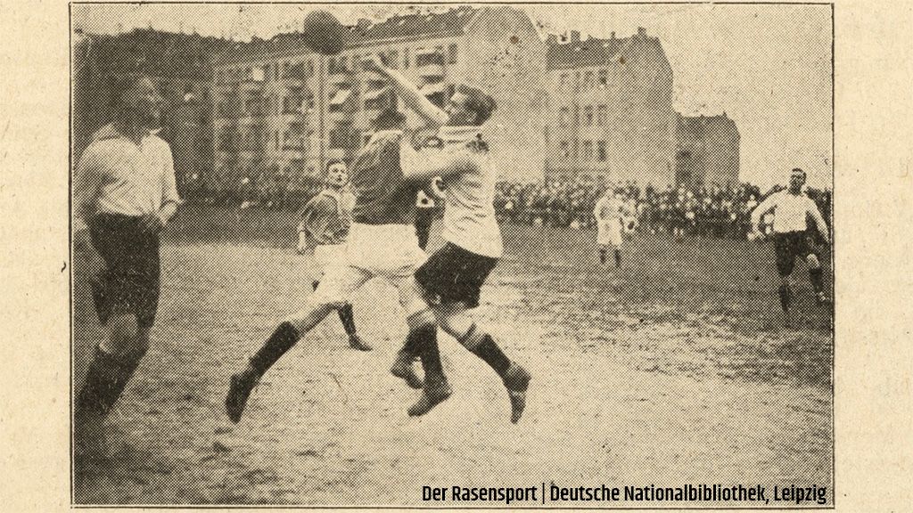 Spielszene im Fußball mit Spielern in historischen Trikots auf einem matschigen Platz, Zuschauer im Hintergrund.