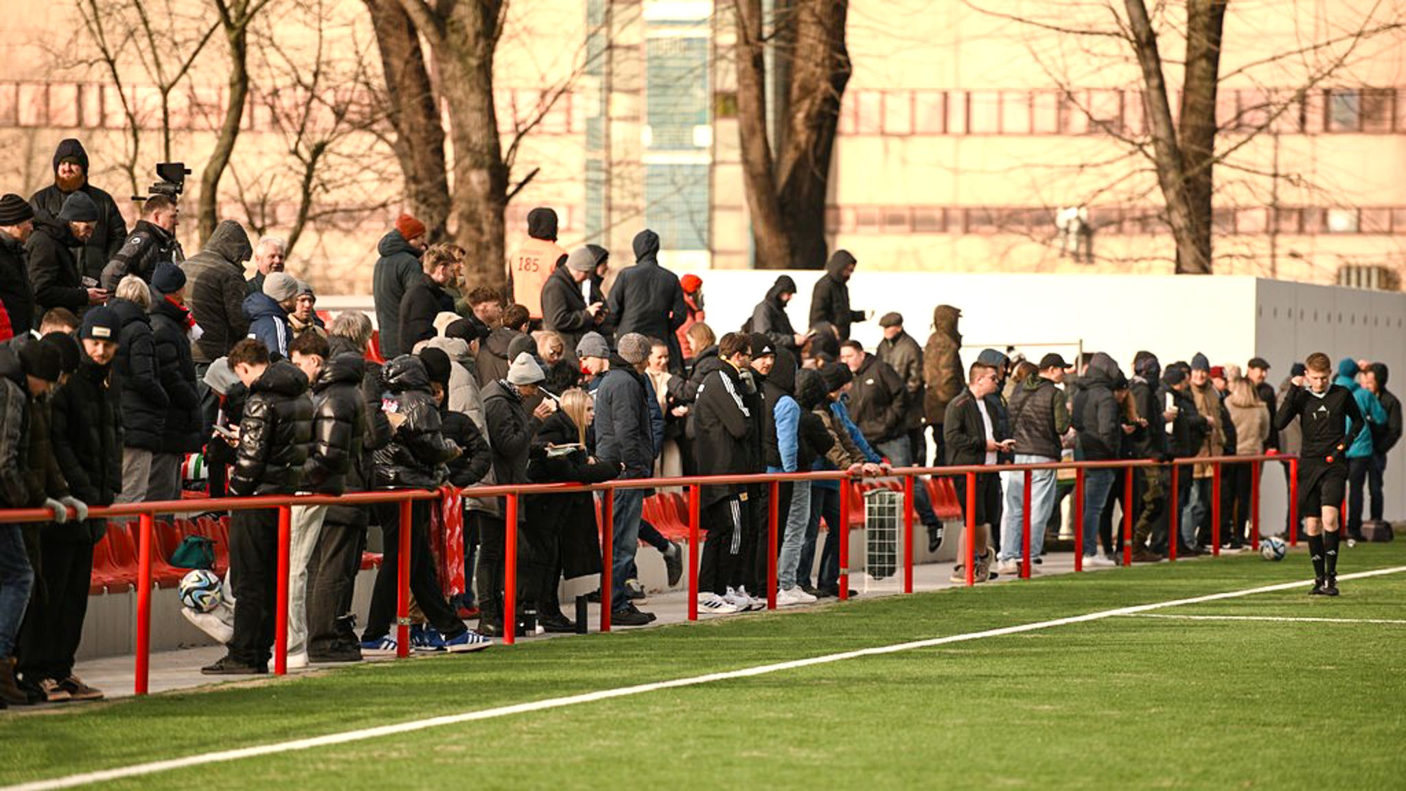 Eine Menschenmenge steht an der Seite eines Fußballfeldes, während ein Spiel stattfindet. Die Zuschauer tragen warme Kleidung.