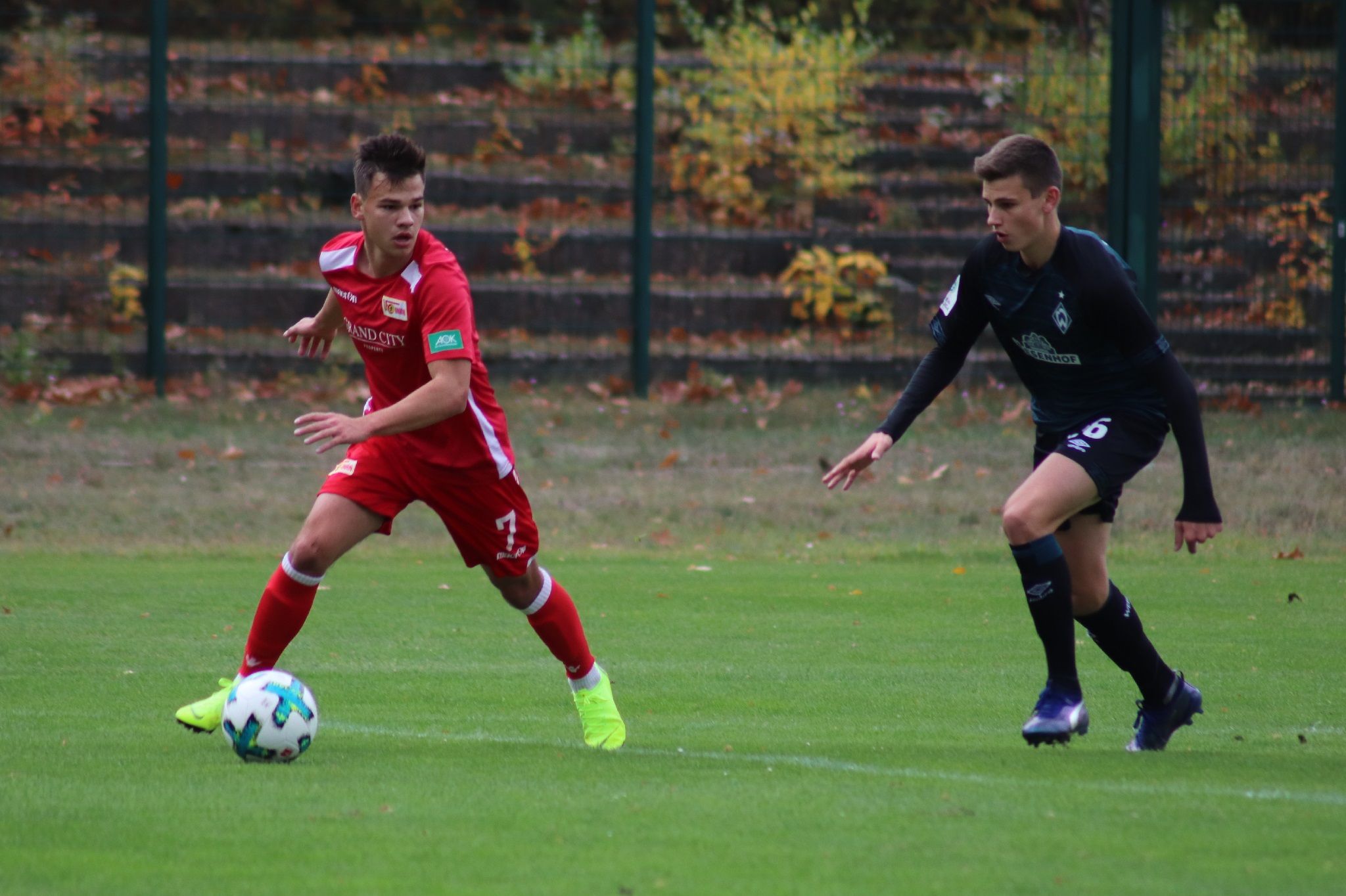 Ein Fußballspieler in rotem Trikot dribbelt den Ball, während ein Gegner in dunklem Trikot Druck ausübt.