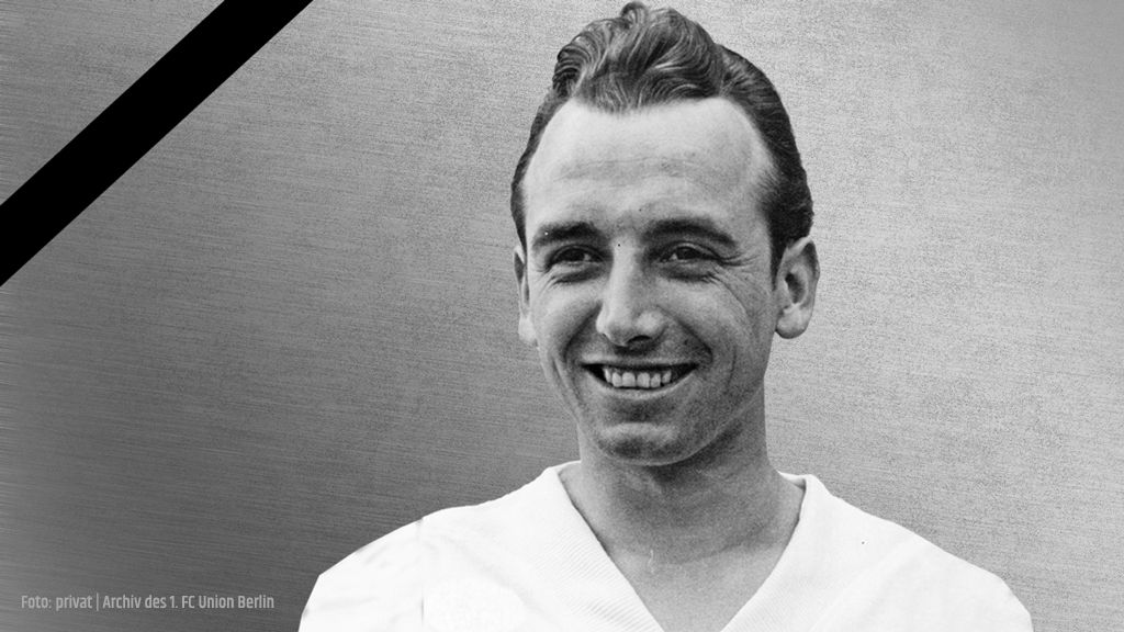 Schwarz-weiße Portraitaufnahme eines lächelnden Mannes in einem oberen Kleidungsstück, vor einem grauen Hintergrund.