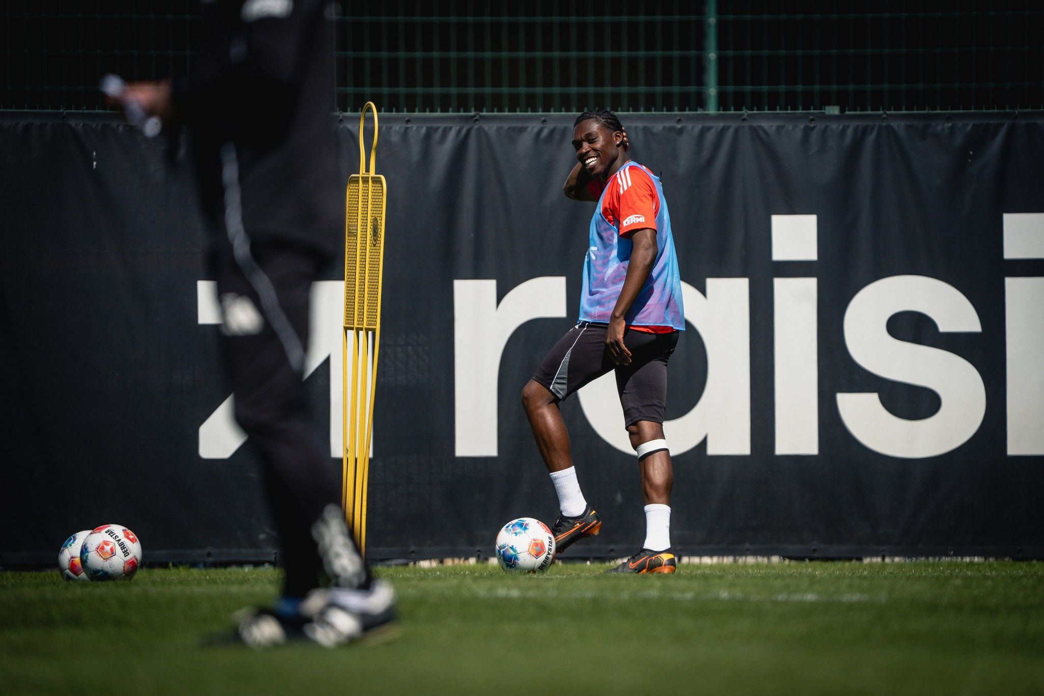 Ein Fußballspieler in Trainingskleidung steht lächelnd auf dem Platz neben einem Markierungsnetz und einem Ball.