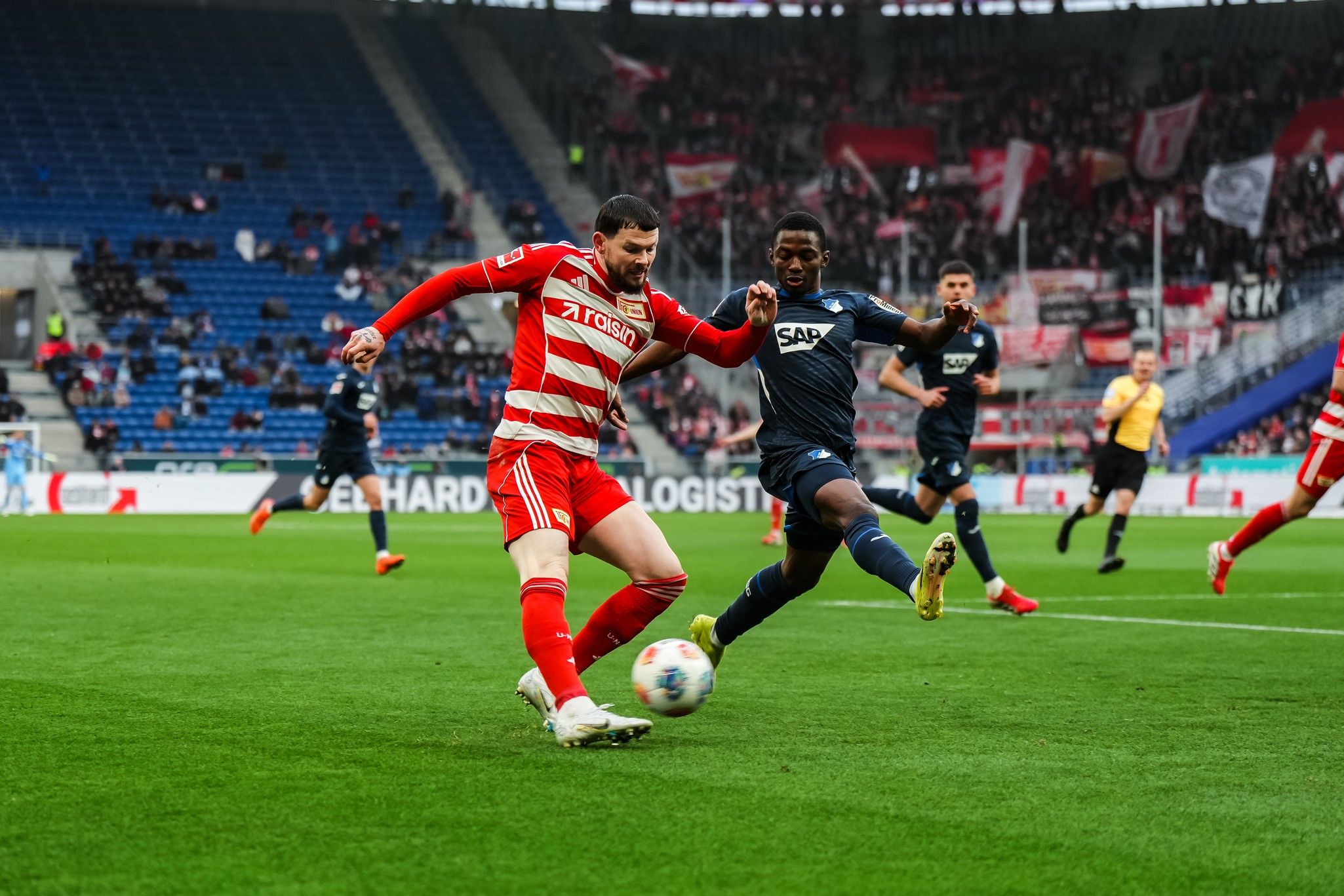 Fußballspiel mit einem Spieler in rotem Trikot, der den Ball vor einem gegnerischen Spieler in blauem Trikot dribbelt.