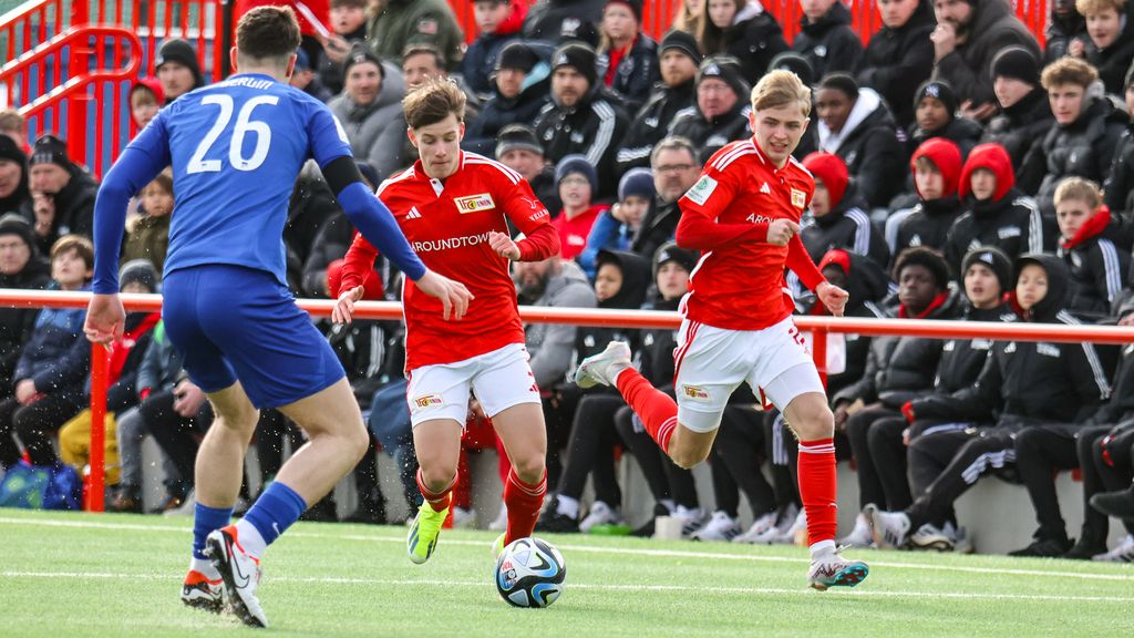 Zwei Spieler in roten Trikots dribbeln den Ball, während ein Spieler in Blau sie verteidigt. Im Hintergrund stehen Zuschauer.