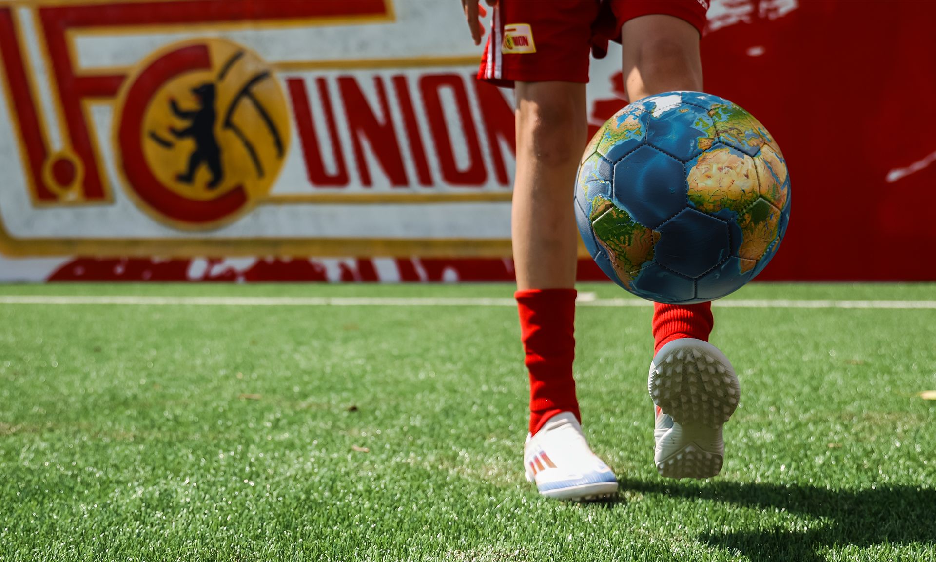 Ein Spieler mit roter Shorts und weißen Schuhen dribbelt einen bunten Fußball auf einem grünen Kunstrasen. Im Hintergrund ein Vereinslogo.