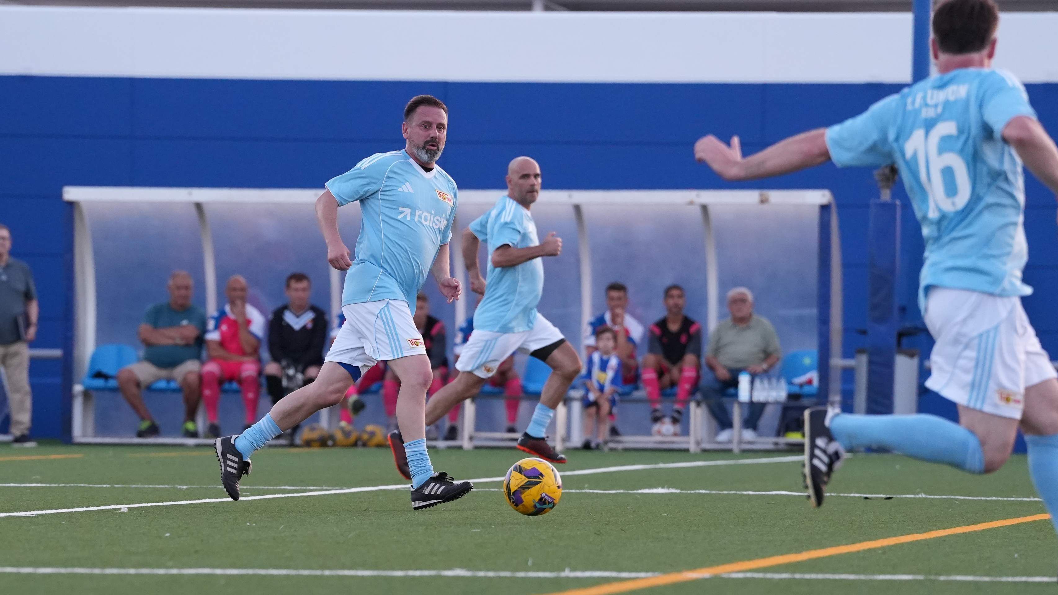Spieler in hellblauen Trikots laufen auf einem Fußballfeld. Im Hintergrund sitzen viele Zuschauer auf Bänken.