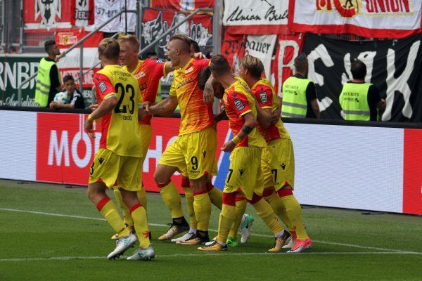 Fußballmannschaft in gelben Trikots jubelt nach einem Tor, während Fans im Hintergrund angeregt feiern.
