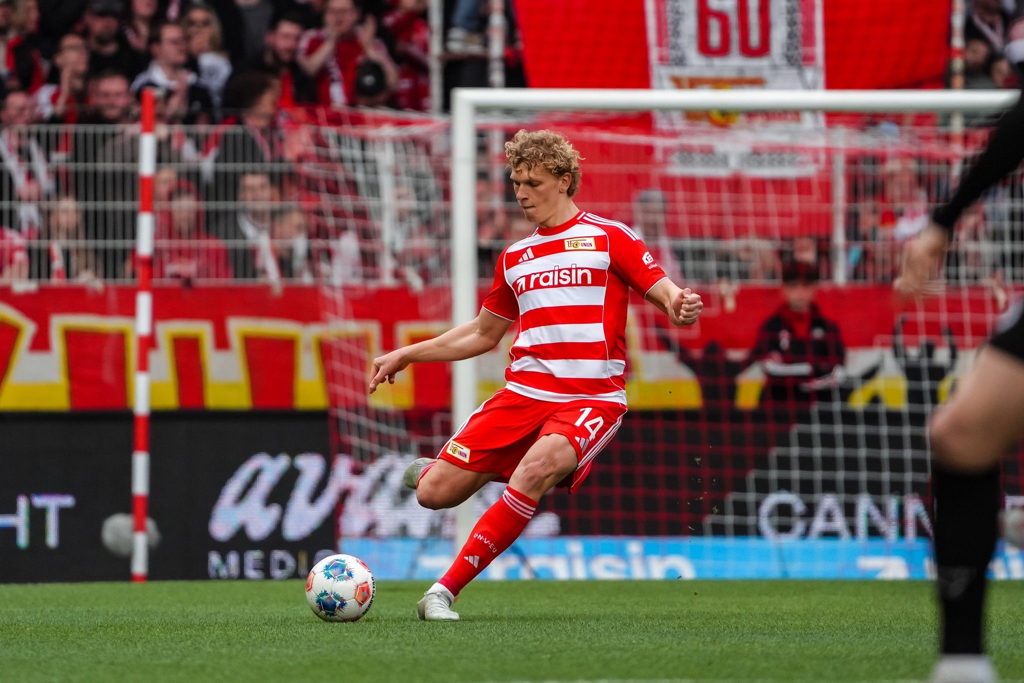 Ein Fußballspieler in rot-weißer Uniform kickt den Ball während eines Spiels, umgeben von jubelnden Fans im Stadion.