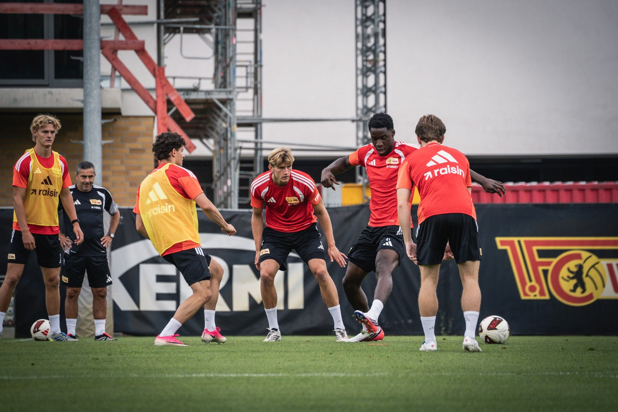 Fußballspieler in roten Trikots trainieren gemeinsam auf dem Feld, während ein Trainer im Hintergrund beobachtet.