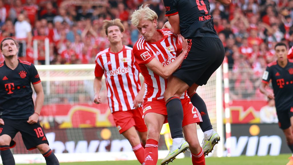 Zwei Fußballspieler in Aktion während eines Spiels, einer führt einen Zweikampf, im Hintergrund jubelnde Fans.