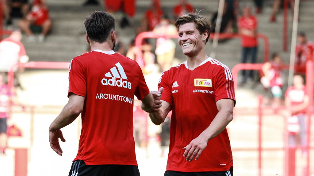 Zwei Fußballspieler in roten Trikots von Union Berlin schütteln sich die Hände und lächeln während eines Spiels.