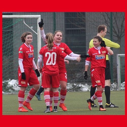 Vier Fußballspielerinnen in roten Trikots feiern einen Torerfolg, während ein Schiedsrichter im Hintergrund steht.