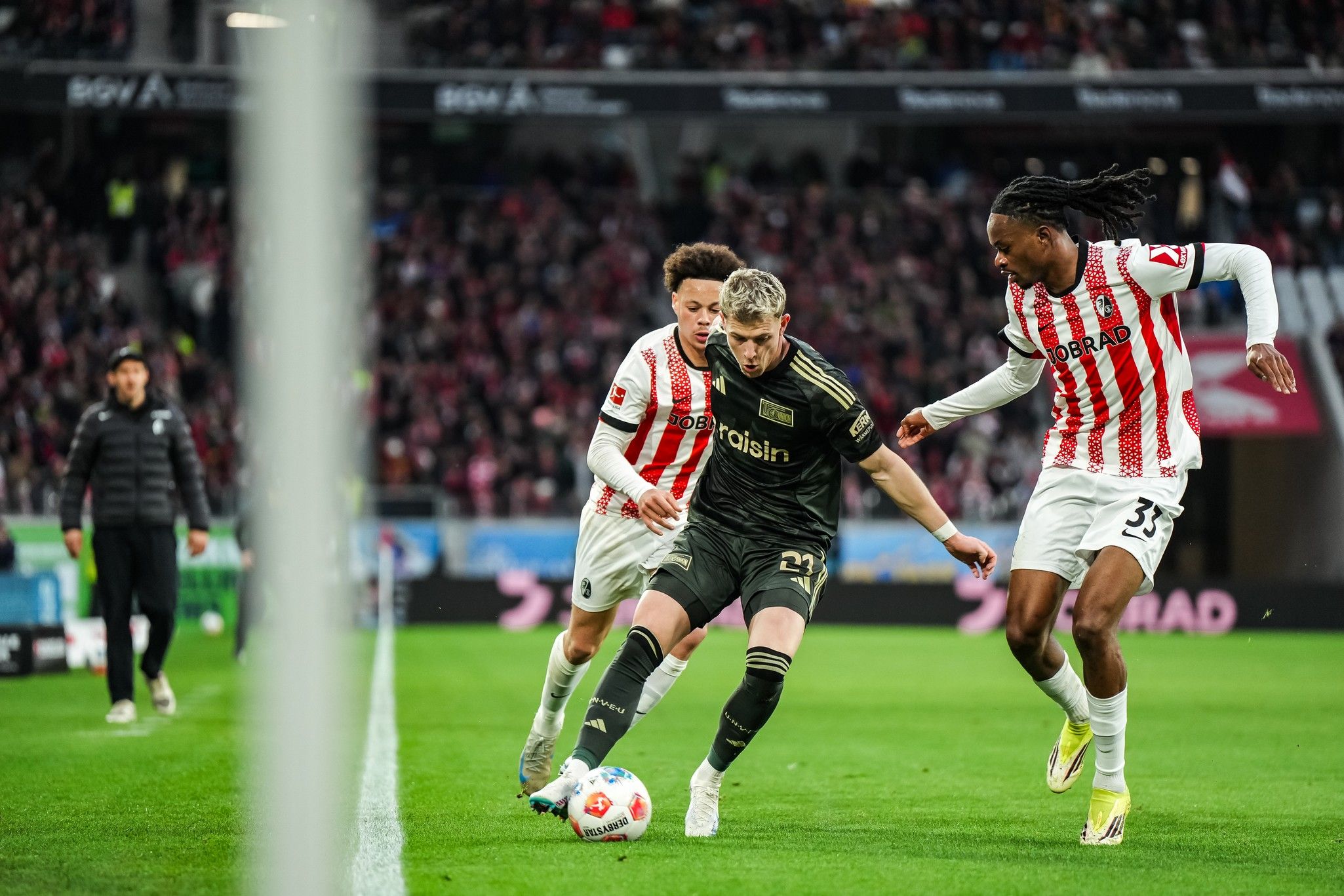 Ein Fußballspieler dribbelt den Ball entlang der Seitenlinie, während zwei Gegner ihn an greifen. Stadion mit Publikum im Hintergrund.