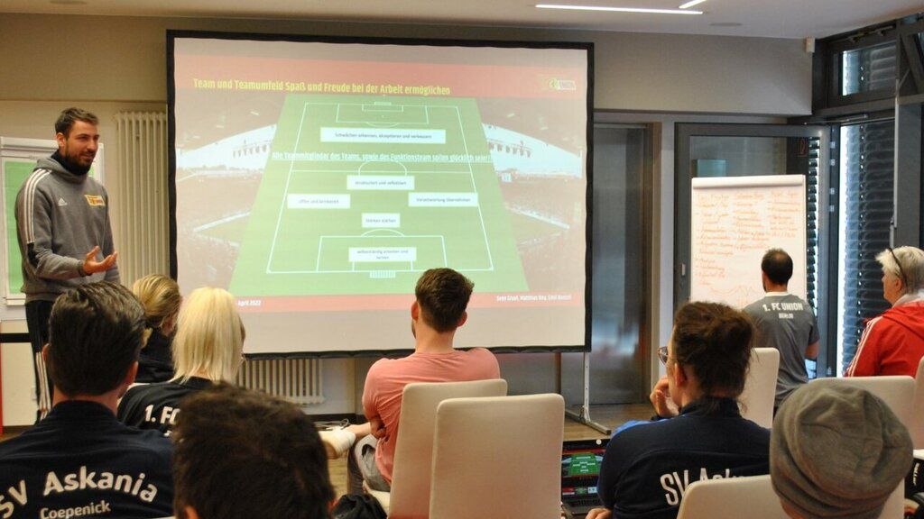 Ein Dozent präsentiert eine taktische Fußballaufstellung vor einer Gruppe von Zuhörern in einem Seminarraum.