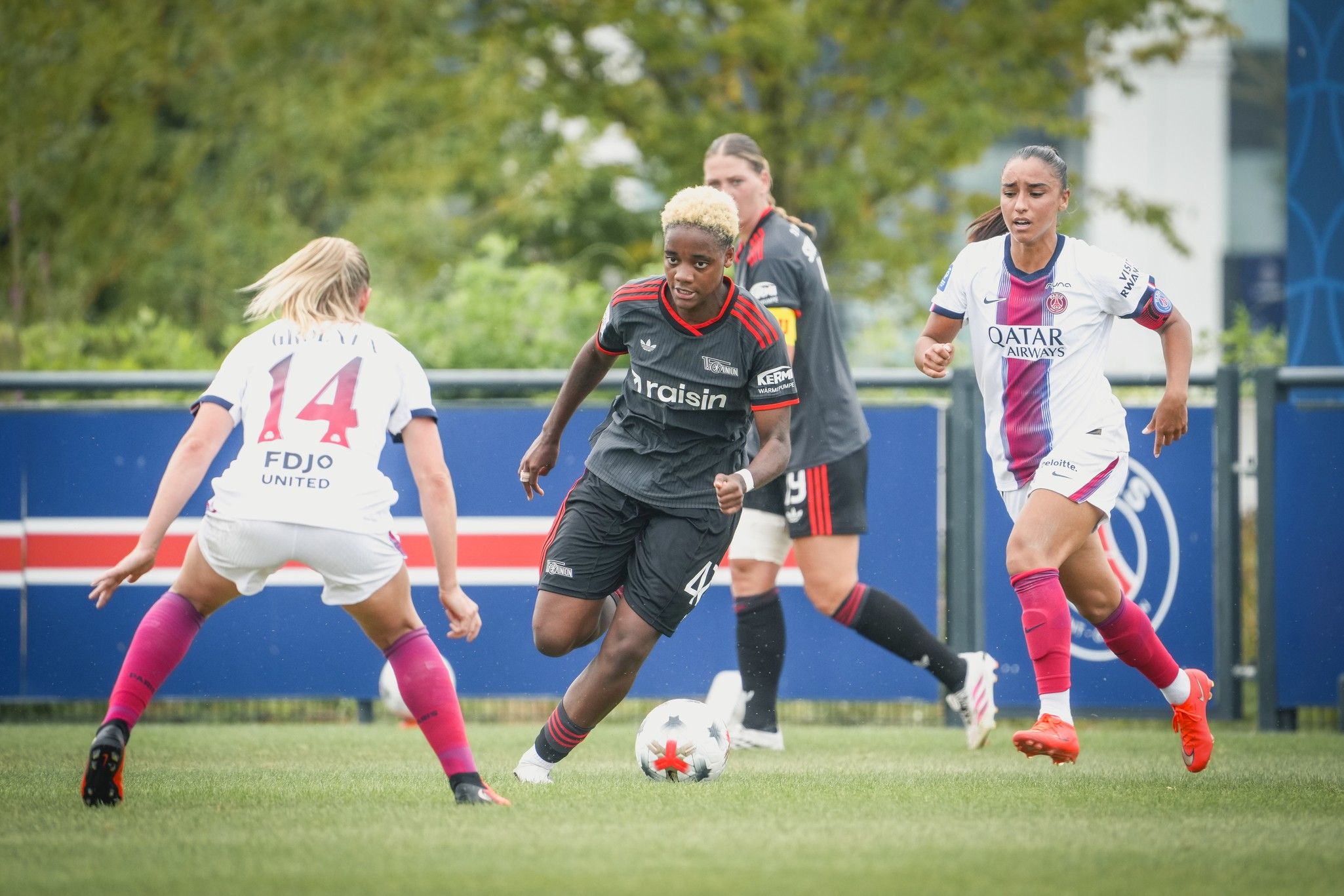 Eine Fußballspielerin im schwarzen Trikot dribbelt den Ball, während zwei Gegnerinnen im weißen und rosa Trikot versuchen, sie zu stoppen.