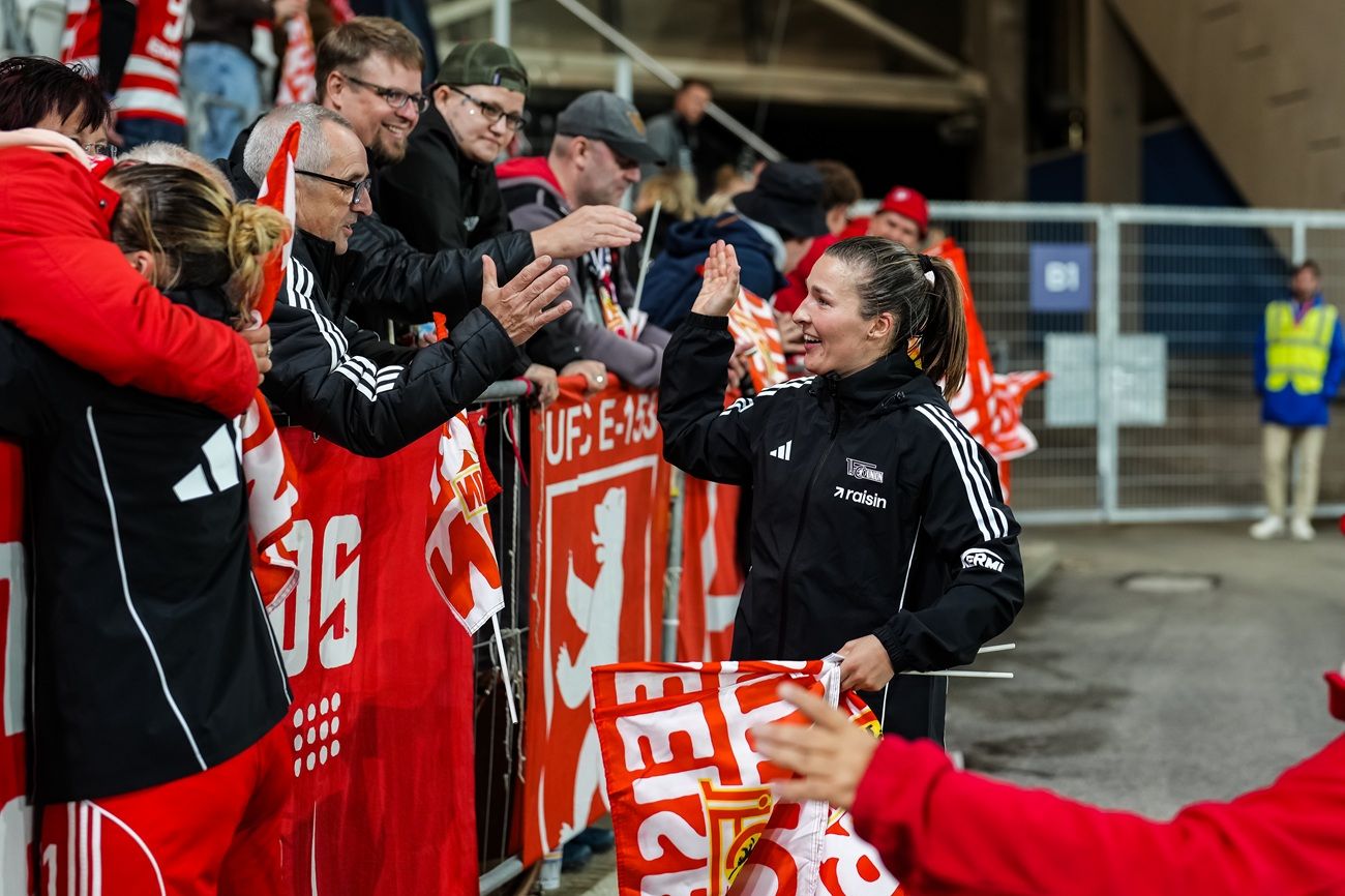 Eine Gruppe von Fans applaudiert und interagiert freudig mit einer Person, die eine Flagge hält, an einem sportlichen Ereignis.
