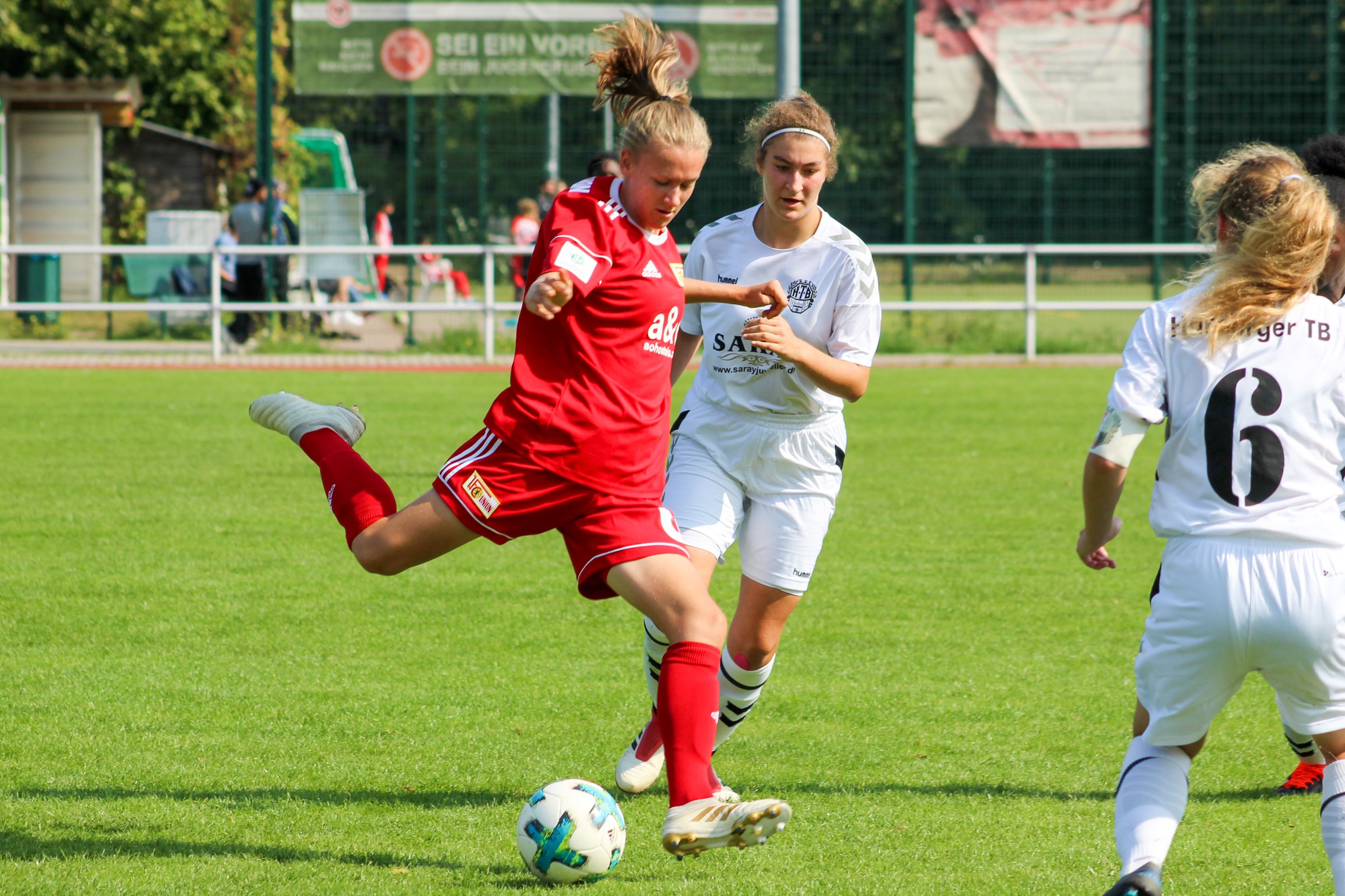 Eine Fußballspielerin in roter Uniform tritt den Ball, während eine Gegnerin in weißer Uniform sie verfolgt.
