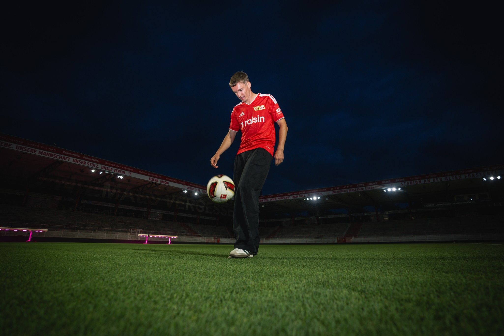 Ein junger Mann in einem roten Fußballtrikot steht auf dem Spielfeld und dribbelt mit einem Fußball bei Nacht.