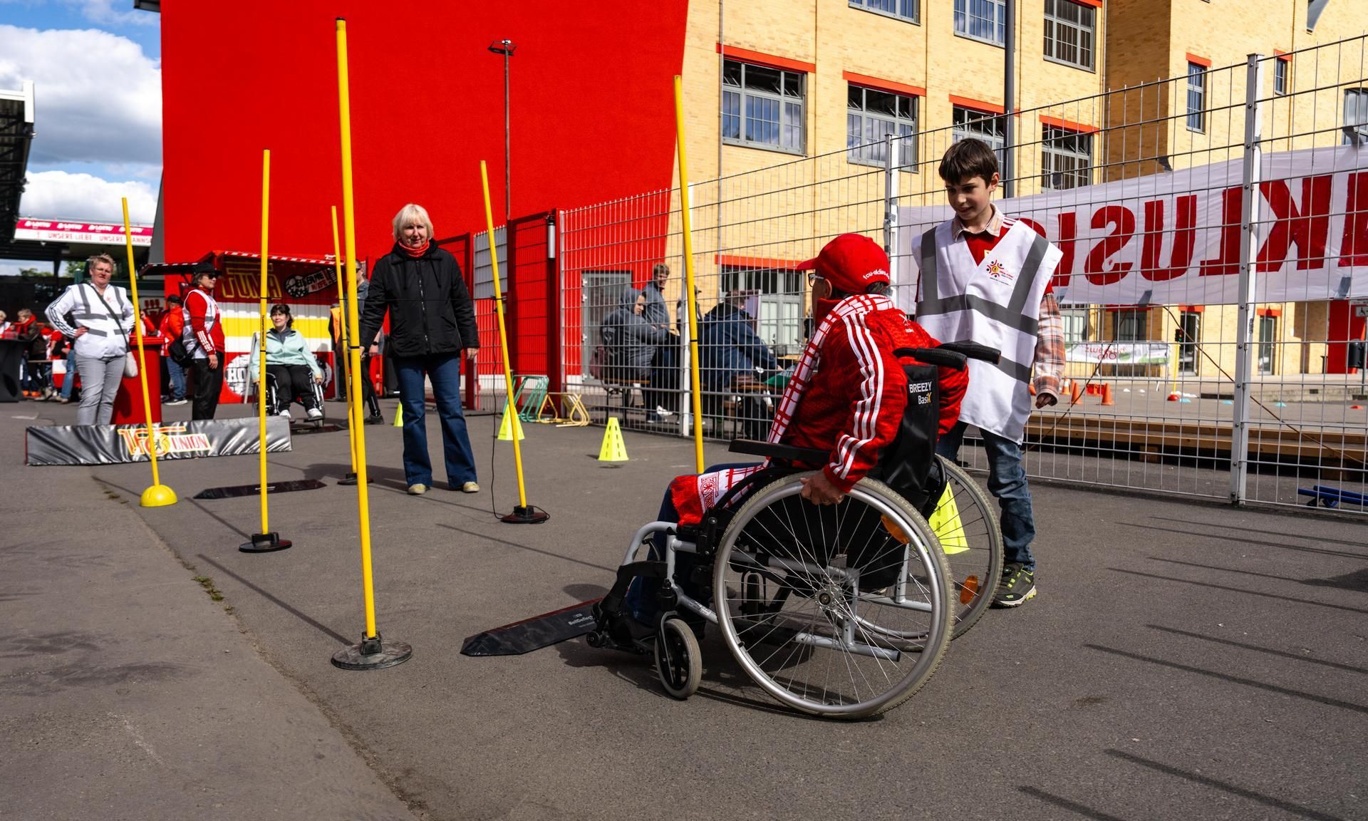Eine Person im Rollstuhl fährt durch einen Parcours mit gelben Stangen, während eine Frau und ein Junge zuschauen.