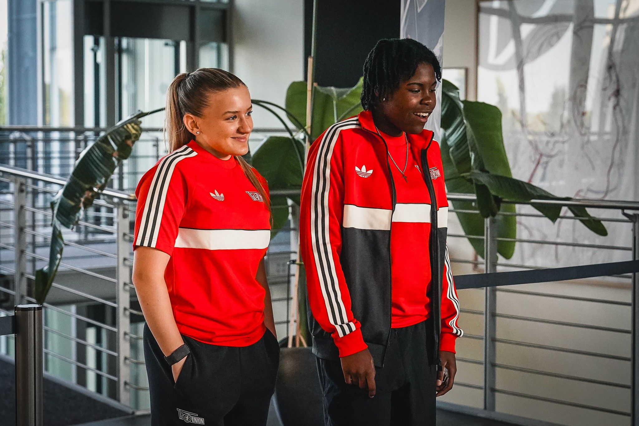 Zwei Frauen in roten Sportanzügen mit weißen und schwarzen Streifen stehen in einem modernen Innenraum und lächeln.