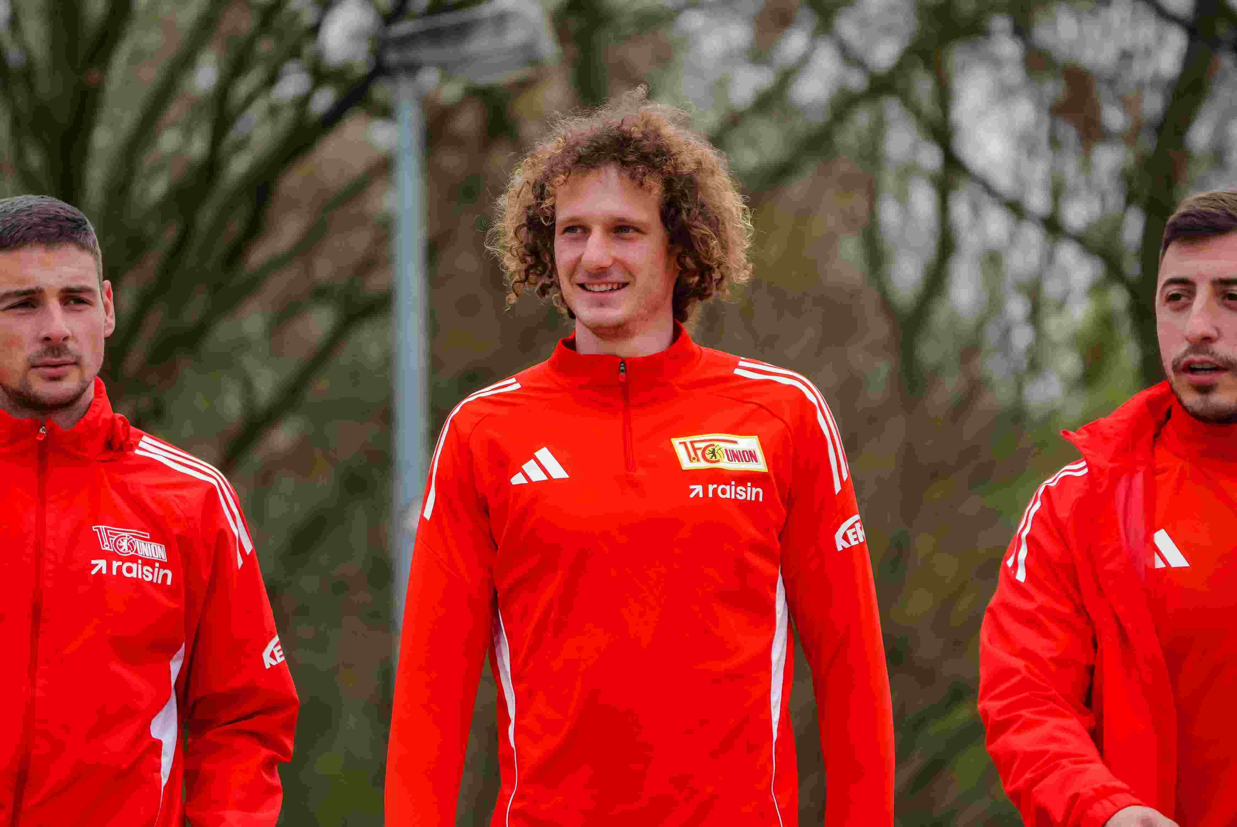 Drei Männer in roten Trainingsjacken stehen zusammen im Freien, lächeln und blicken leicht nach rechts.